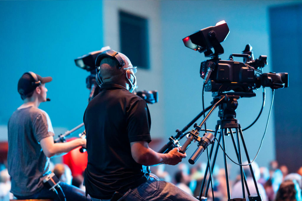 Two cameramen operate professional video cameras at an indoor event with a lively audience.